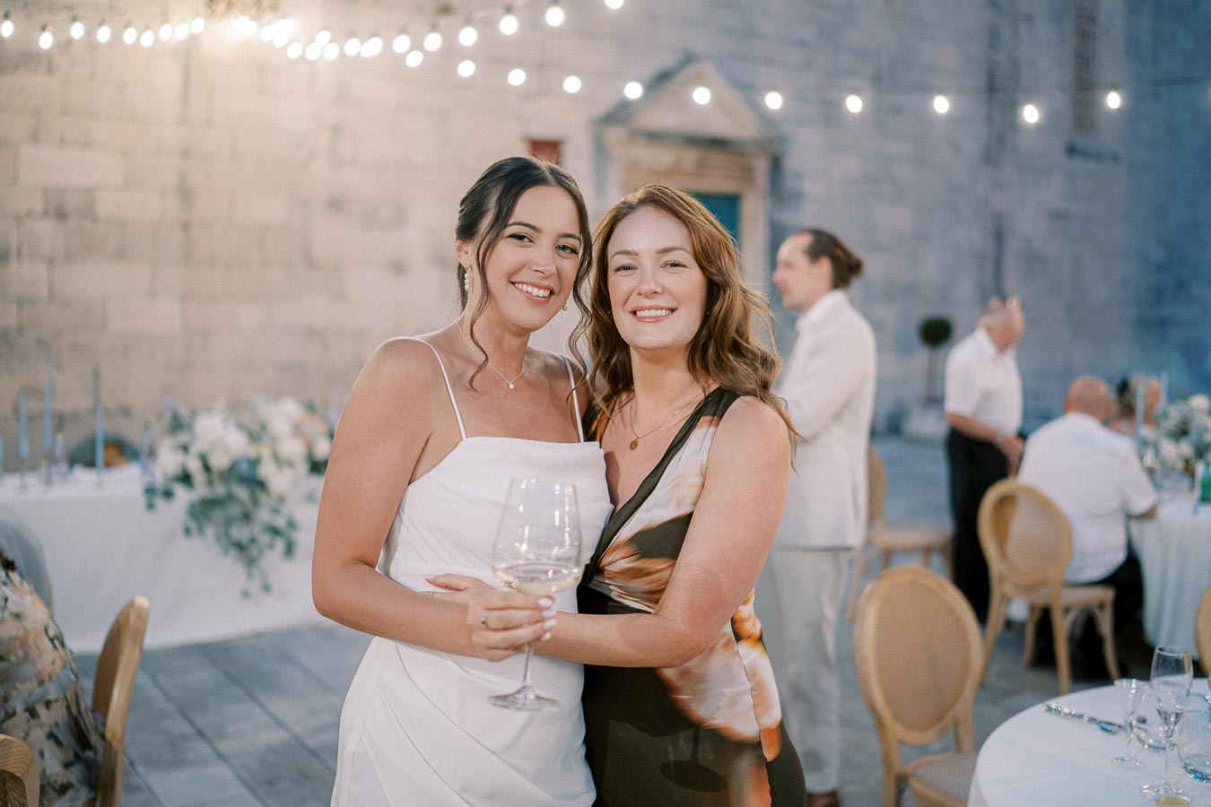Two women smiling and holding a wine glass at an elegant outdoor event with string lights and a stone wall background.