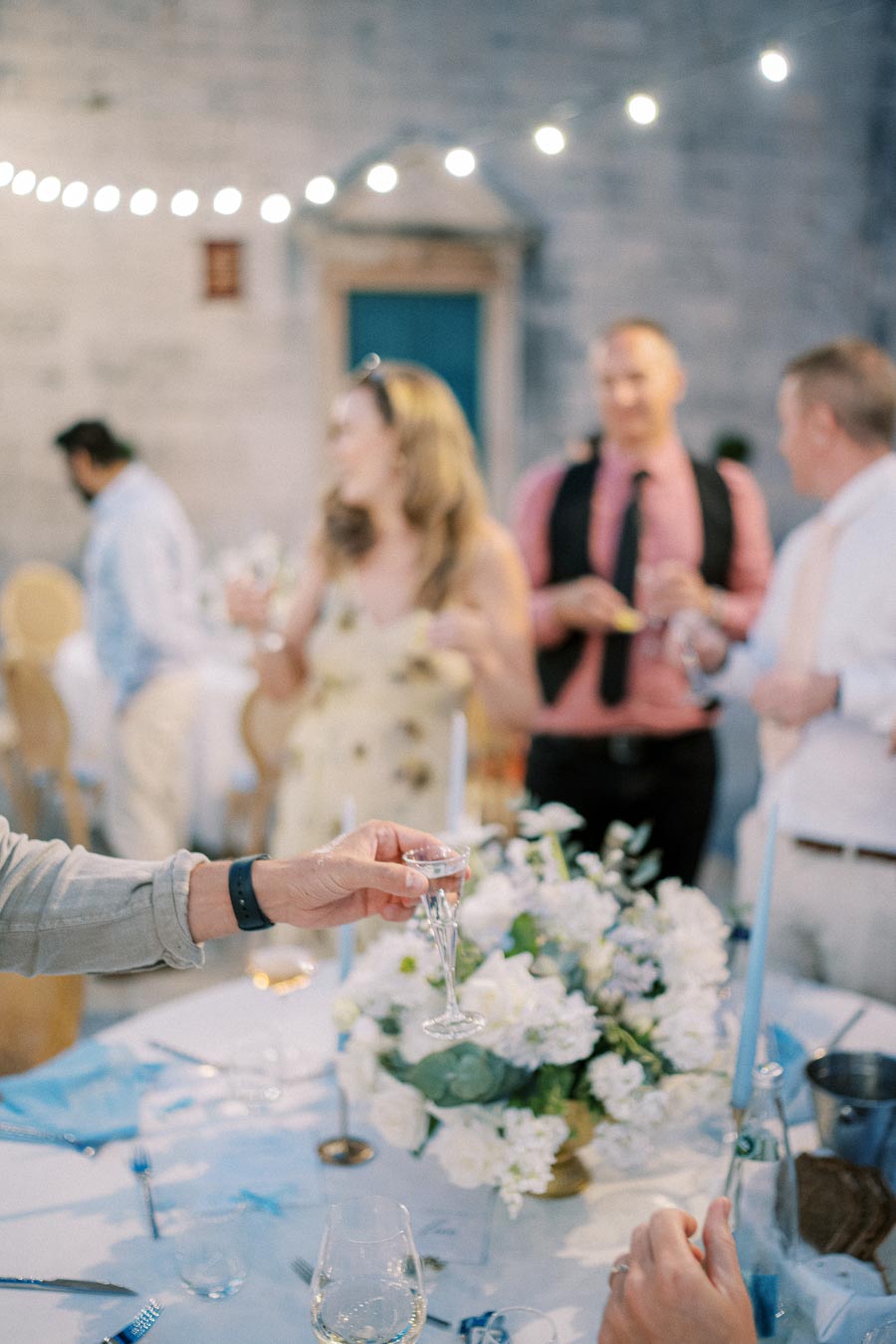A group of people socializing at an elegant event, with a focus on a hand holding a champagne flute over a beautifully decorated table with white flowers and blue accents, soft lighting creating a warm atmosphere.
