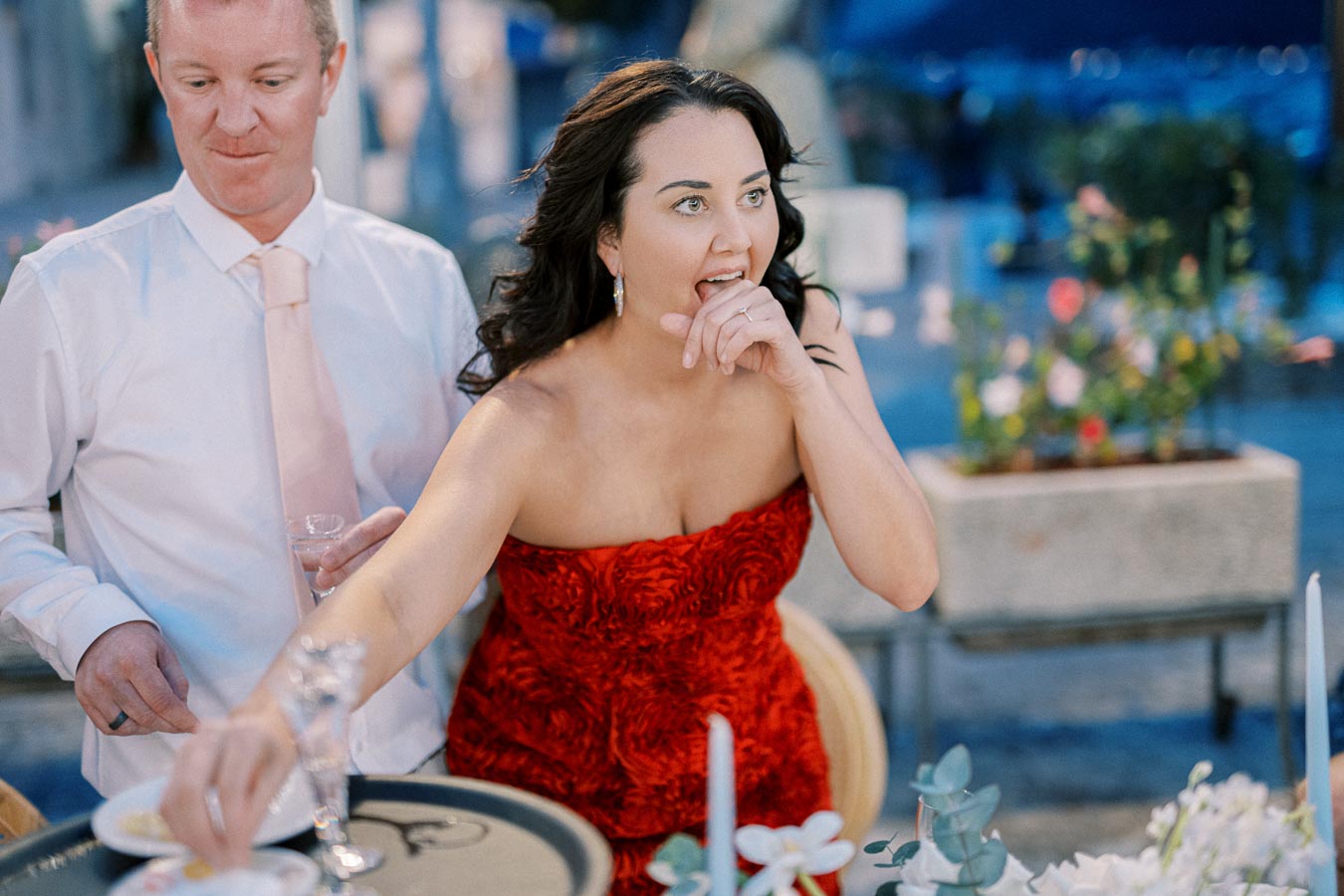 A woman in a strapless red dress enjoying an outdoor event with a man in a white shirt and pink tie, playfully reaching for items on a table amidst a lively atmosphere.