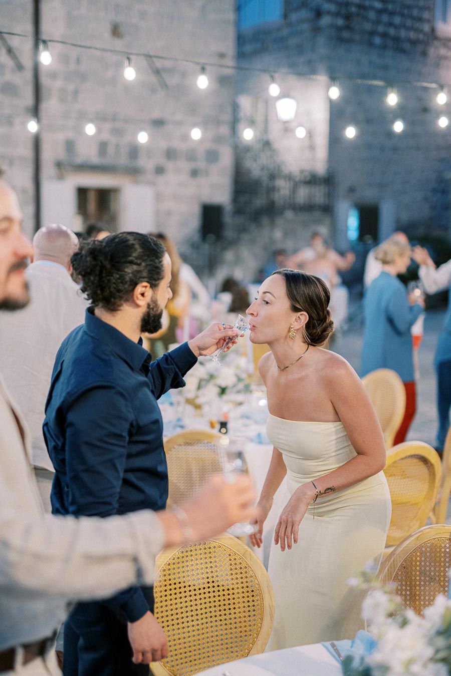 A couple sharing a drink during a festive outdoor wedding reception, with string lights illuminating the evening setting. Guests are seated at decorated tables in the background, adding to the joyous atmosphere.