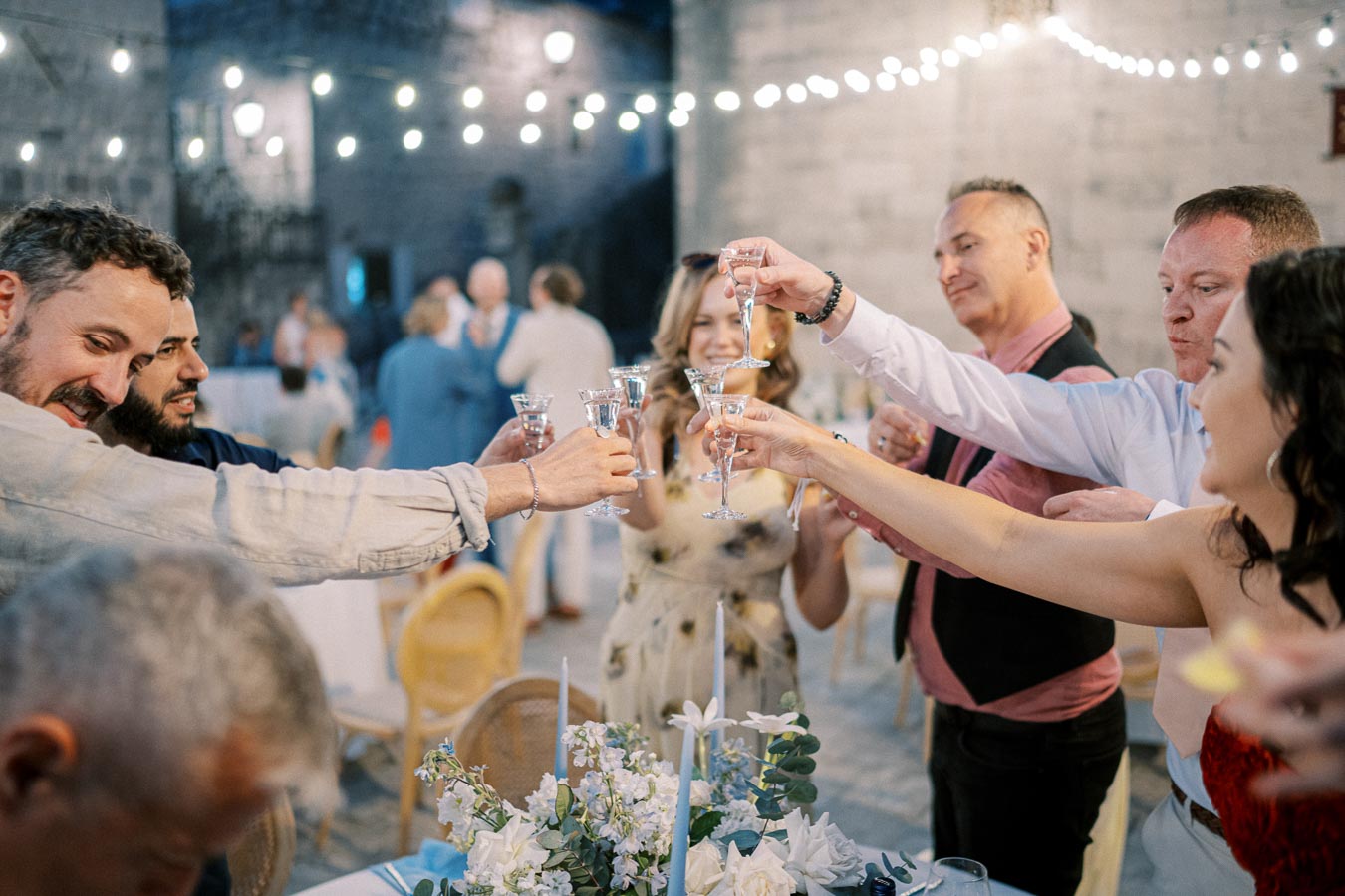 A group of people clinking glasses in a celebratory toast at an outdoor evening event, with string lights and a floral centerpiece creating a festive atmosphere.