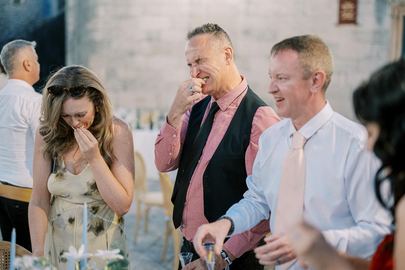 A group of people enjoying a moment at a social event, with a man in a pink shirt and vest reacting humorously while eating, a woman beside him smiling, and another man in a white shirt laughing. The setting appears to be a cheerful gathering with decorative elements, suggesting a festive atmosphere.