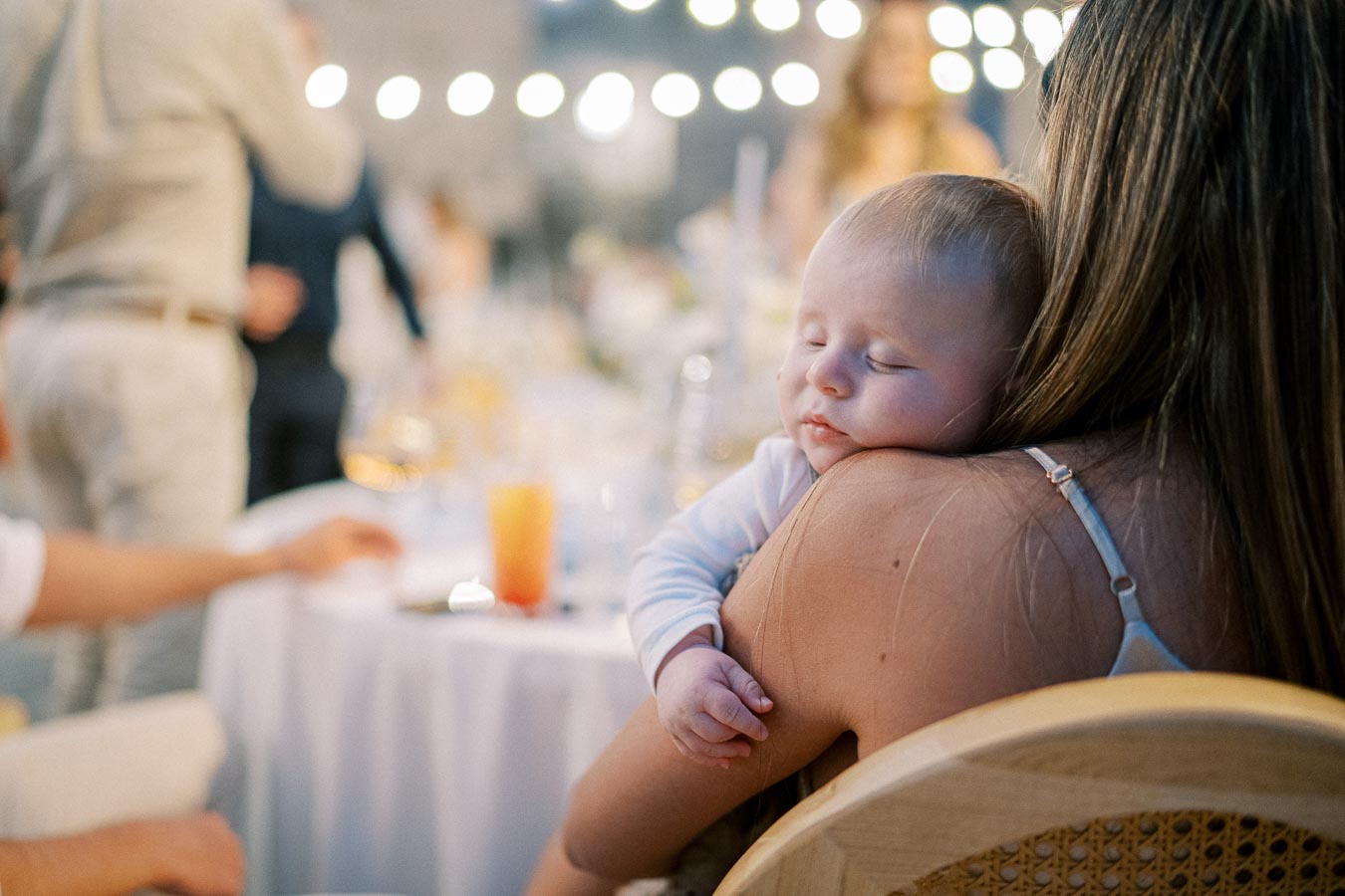 A peaceful baby sleeping on a woman's shoulder at a wedding reception, with soft ambient lighting and blurred background.