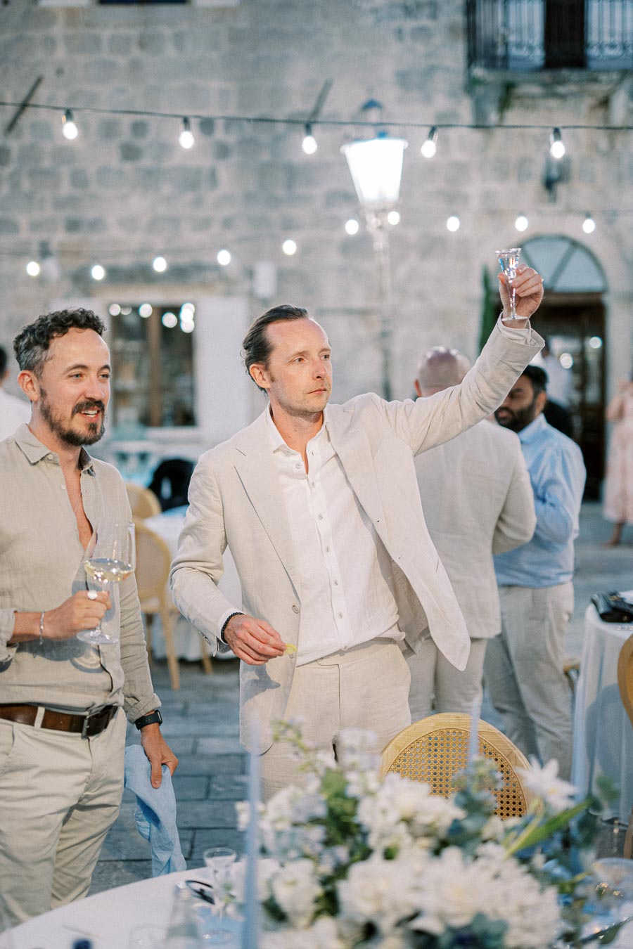 Two men in light-colored suits toast with wine glasses at an elegant, outdoor event with string lights and floral table setting.