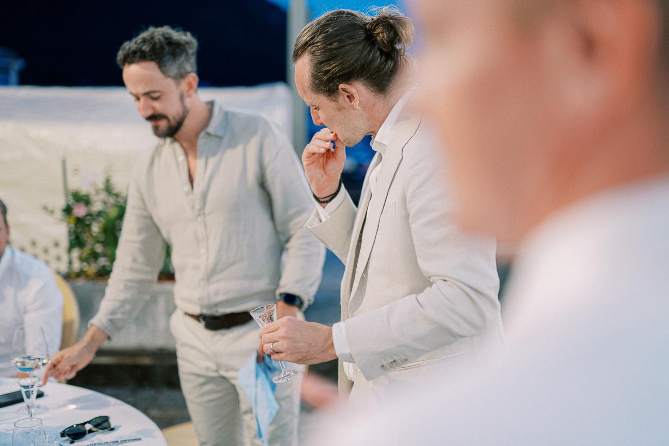 Two men in light-colored suits enjoying a social gathering with drinks, set in an outdoor environment with a relaxed atmosphere.