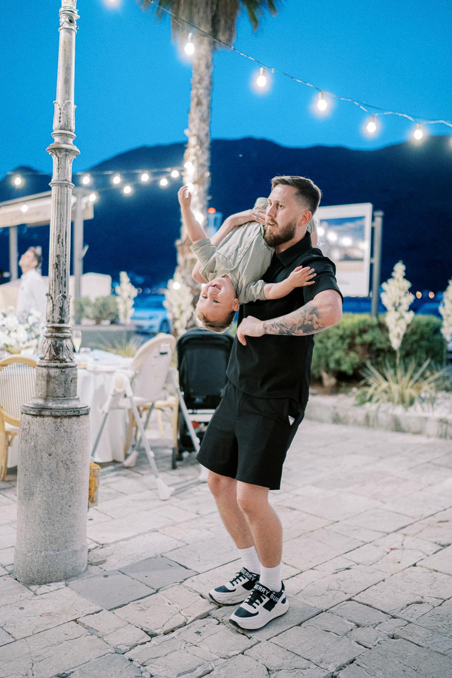 A man playfully swings a child upside down on a tiled outdoor patio with string lights overhead, surrounded by tables and a stroller, against a backdrop of mountains and a clear evening sky.