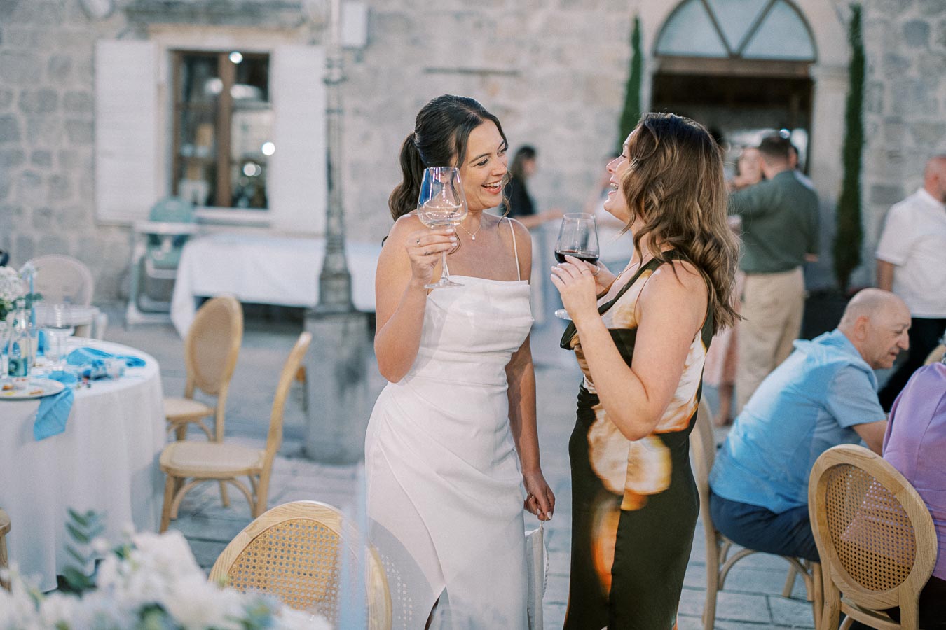 Two women in elegant dresses holding wine glasses and laughing at an outdoor wedding reception, with tables and guests in the background.