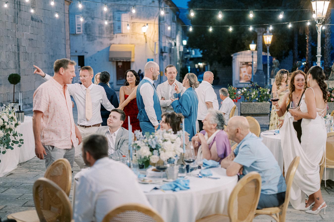 Outdoor evening celebration with people socializing around tables adorned with floral arrangements and string lights, creating a festive and elegant atmosphere.