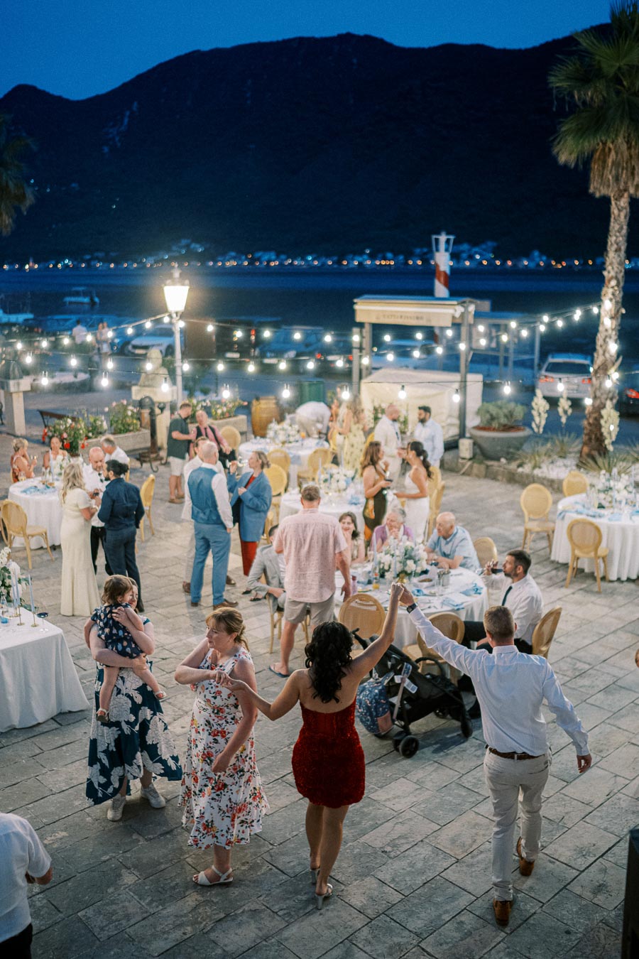 Outdoor evening wedding reception with guests dancing and socializing near beautifully decorated tables, set against a scenic backdrop of mountains and string lights.