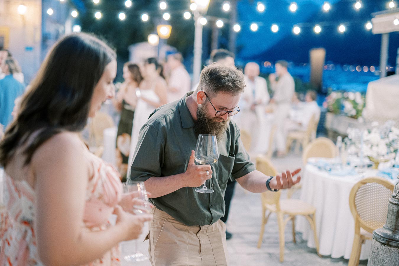 A man with a beard, wearing glasses, enjoys a lively evening outdoor event, holding a wine glass and gesturing playfully, surrounded by groups of people mingling under string lights.