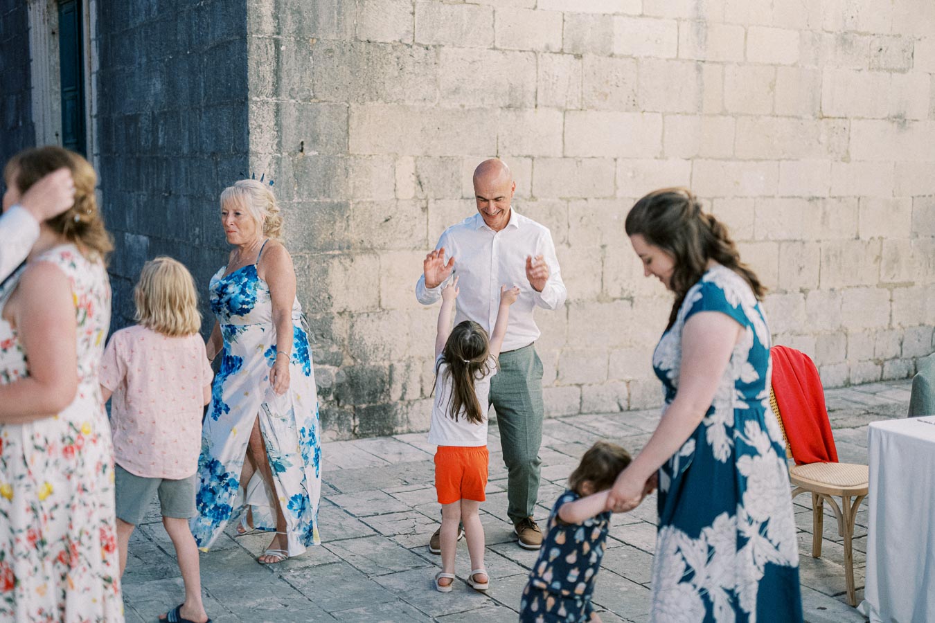 A joyful family gathering with people dancing outdoors on a stone pavement. Adults and children are enjoying quality time together near a historic stone wall, dressed in colorful summer outfits.
