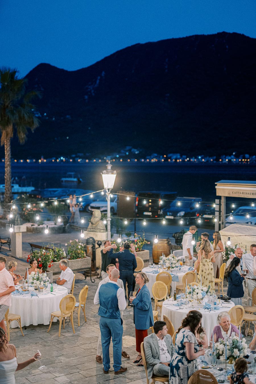 Outdoor evening wedding reception by the sea with guests socializing around elegantly decorated tables under string lights, with a mountainous backdrop and palm trees.