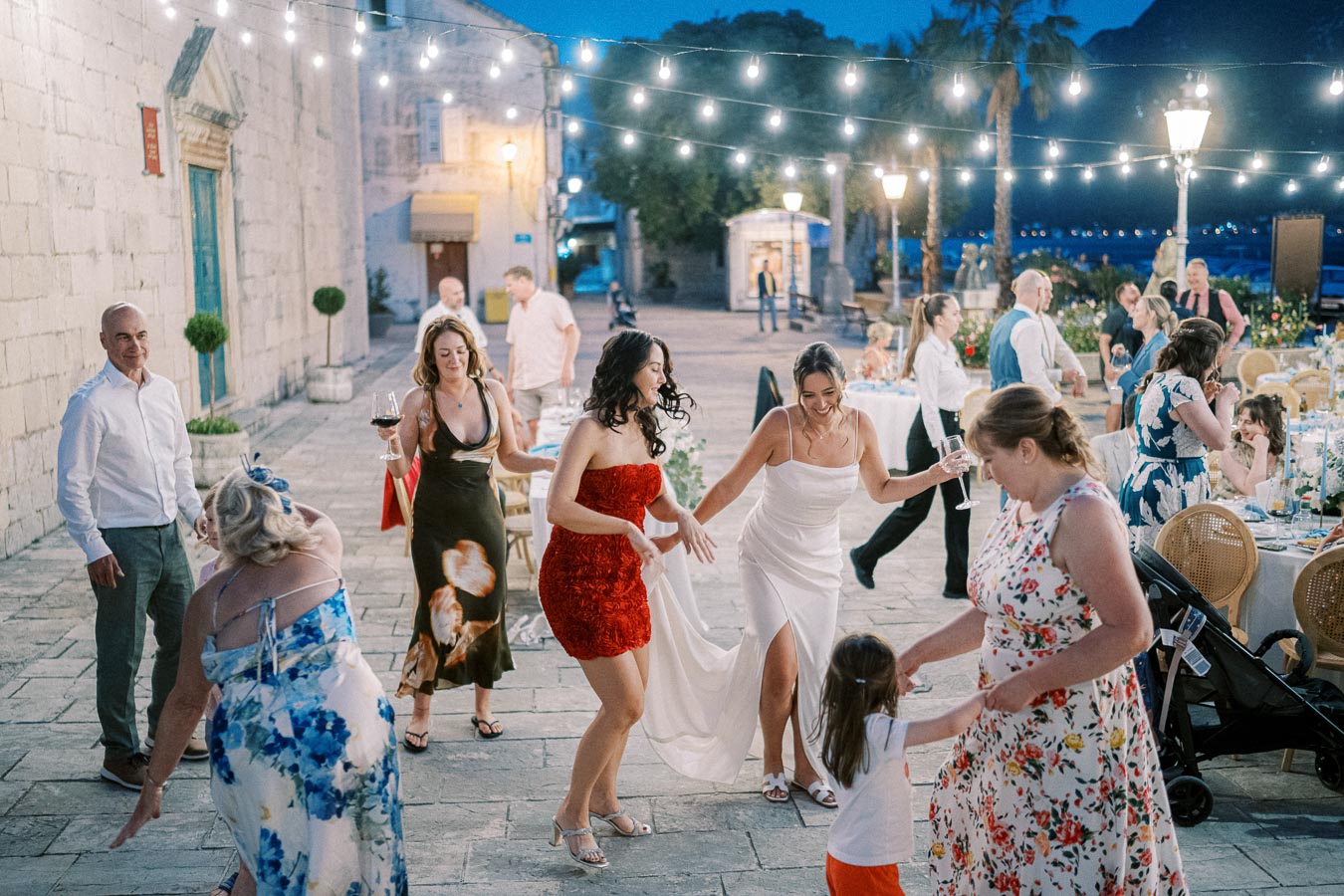 A lively outdoor wedding reception with guests dancing under string lights. The scene captures the joy and celebration, featuring elegantly dressed individuals enjoying the evening on a stone patio, set against a backdrop of rustic architecture and decorative lighting.