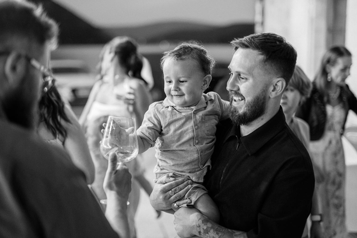 A black and white photo of a joyful man holding a smiling toddler while toasting with another adult at a social gathering. The background features blurred people and a scenic outdoor setting, creating a warm and festive atmosphere.