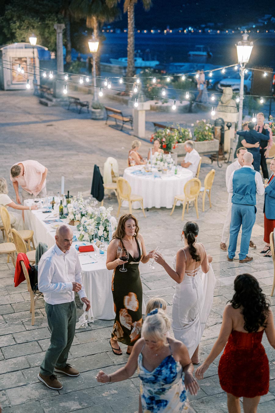 Outdoor evening wedding reception with guests dancing and dining under string lights, with elegantly set tables and scenic waterfront views in the background.