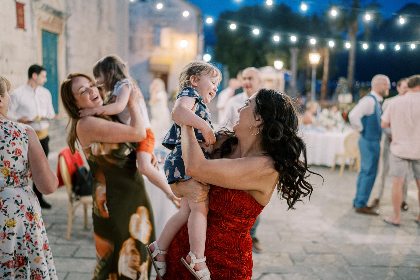 A lively outdoor wedding reception with guests dancing and laughing under string lights, featuring a woman in a red dress joyfully lifting a child.