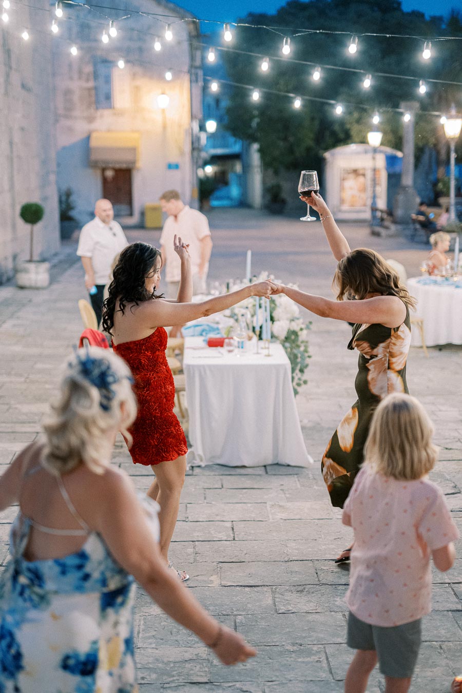 People enjoying an outdoor evening event, with string lights above in a picturesque courtyard. A woman in a red dress and another in a patterned dress dance joyfully, one holding a glass of wine. Others, including a child, watch and participate in the festivities, creating a lively and cheerful atmosphere.