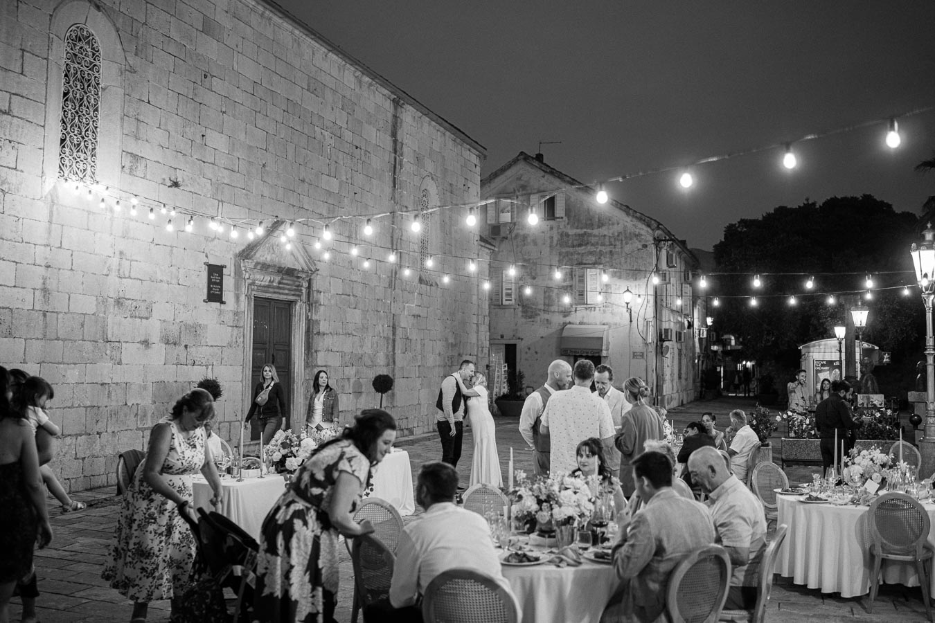 Black and white outdoor wedding reception scene at night, featuring a bride and groom embracing under string lights, surrounded by guests seated at elegantly decorated tables in a historic courtyard.