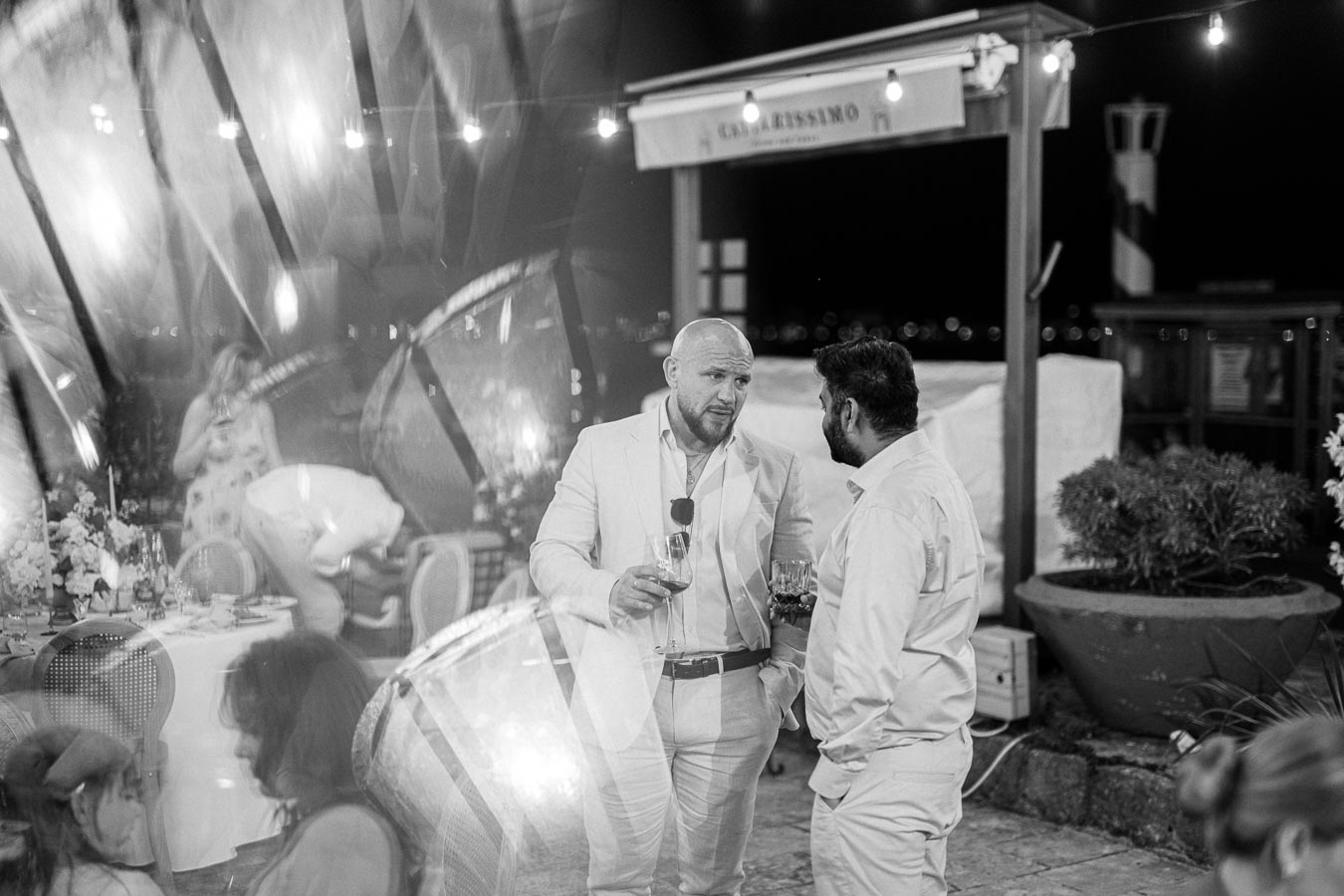 Black and white photo of two men in suits conversing at an elegant outdoor event, holding drinks and standing near a table set with flowers and glassware, with decorative lights and blurred guests in the background.