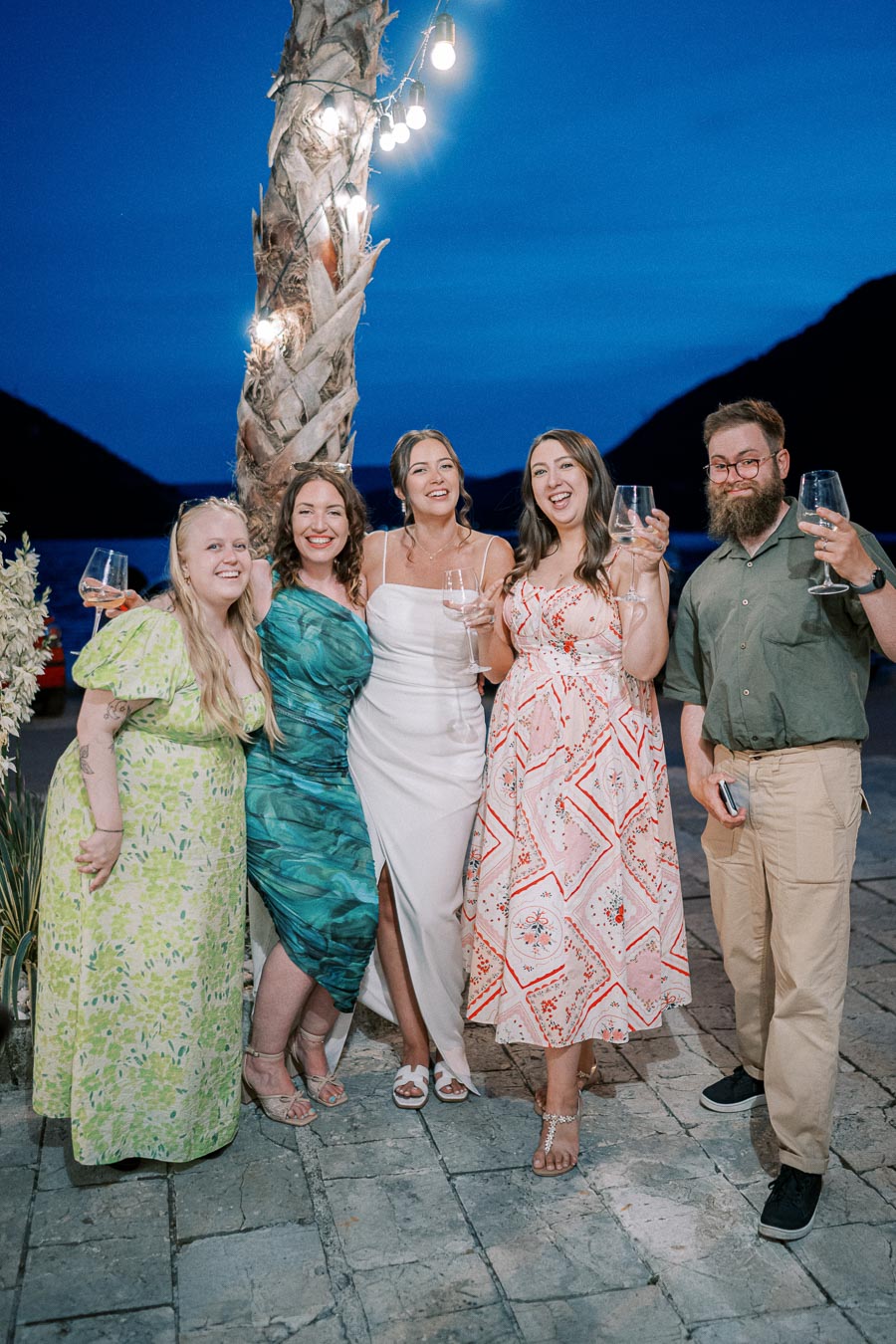 A group of five people smiling and holding wine glasses, standing outdoors at night near a palm tree with string lights, dressed in colorful summer attire against a backdrop of dark blue skies and mountains.