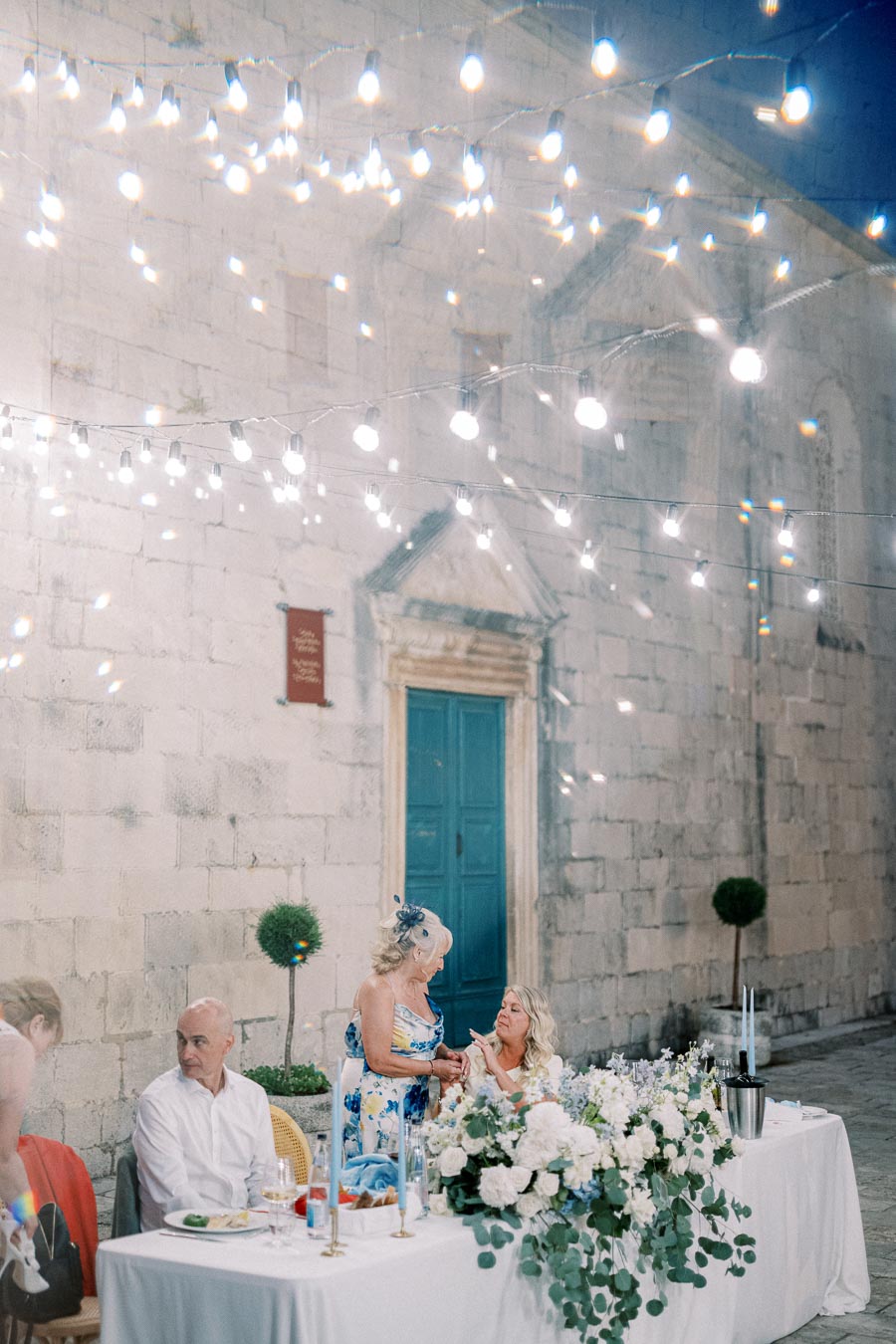 Outdoor wedding reception scene with elegant table setting, floral centerpieces, and guests conversing under twinkling string lights, in front of a historical stone building with a blue door.
