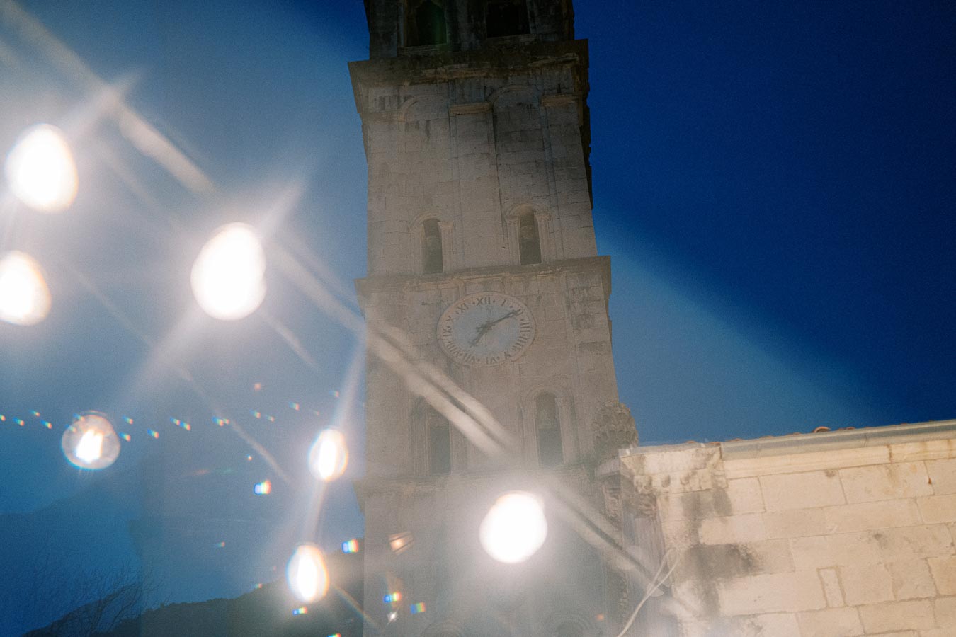 Historic stone clock tower with Roman numerals, illuminated against a clear night sky, with glowing light orbs in the foreground, creating a dreamy atmosphere.