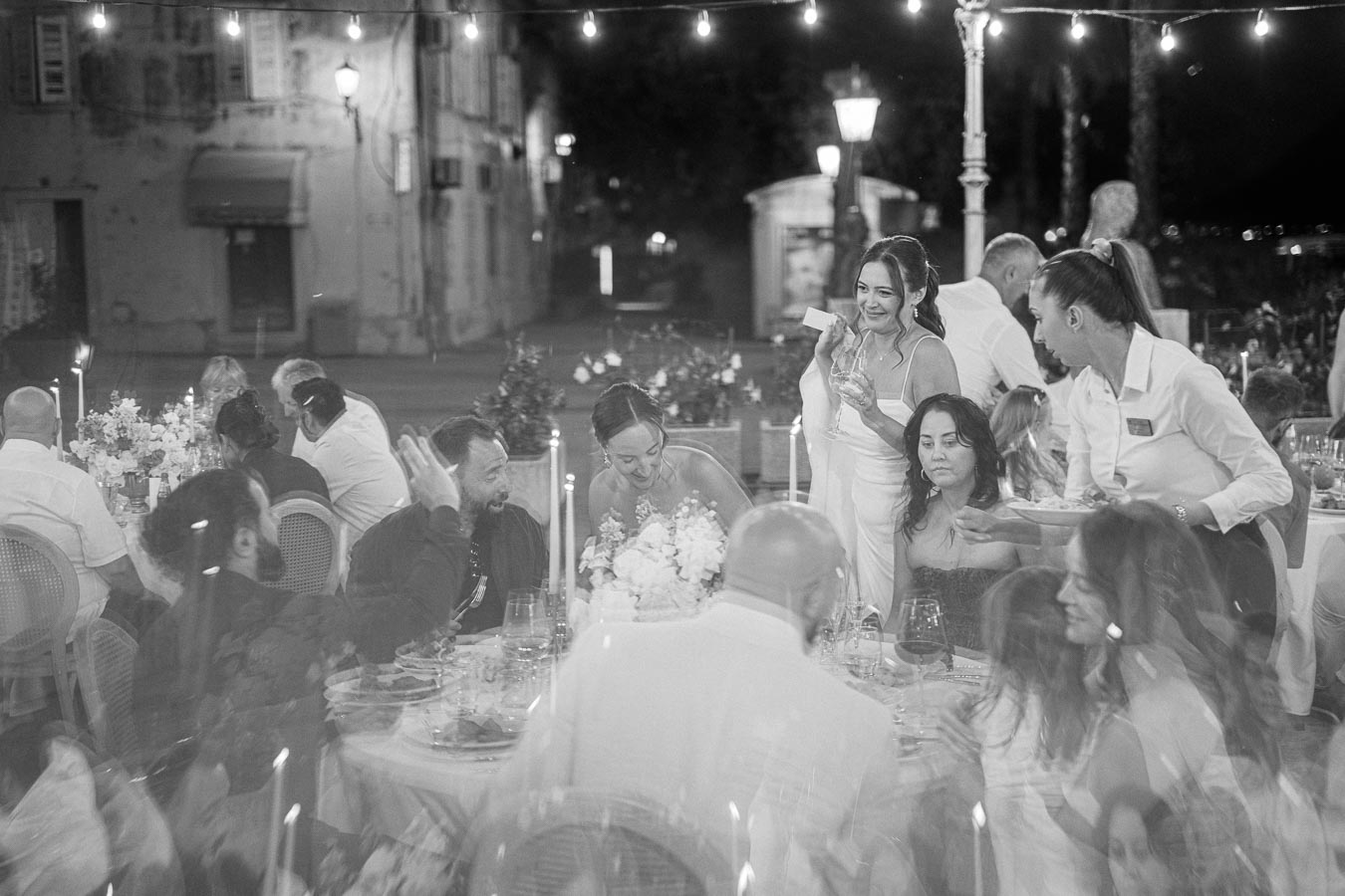 A lively outdoor wedding reception at night with guests enjoying food and conversation under string lights. A bride engages with attendees at the table, creating a warm and festive atmosphere, with a server attending to the guests.
