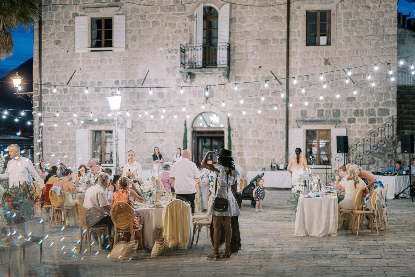 Outdoor wedding reception at night with guests dining under string lights, set against a rustic stone building backdrop.