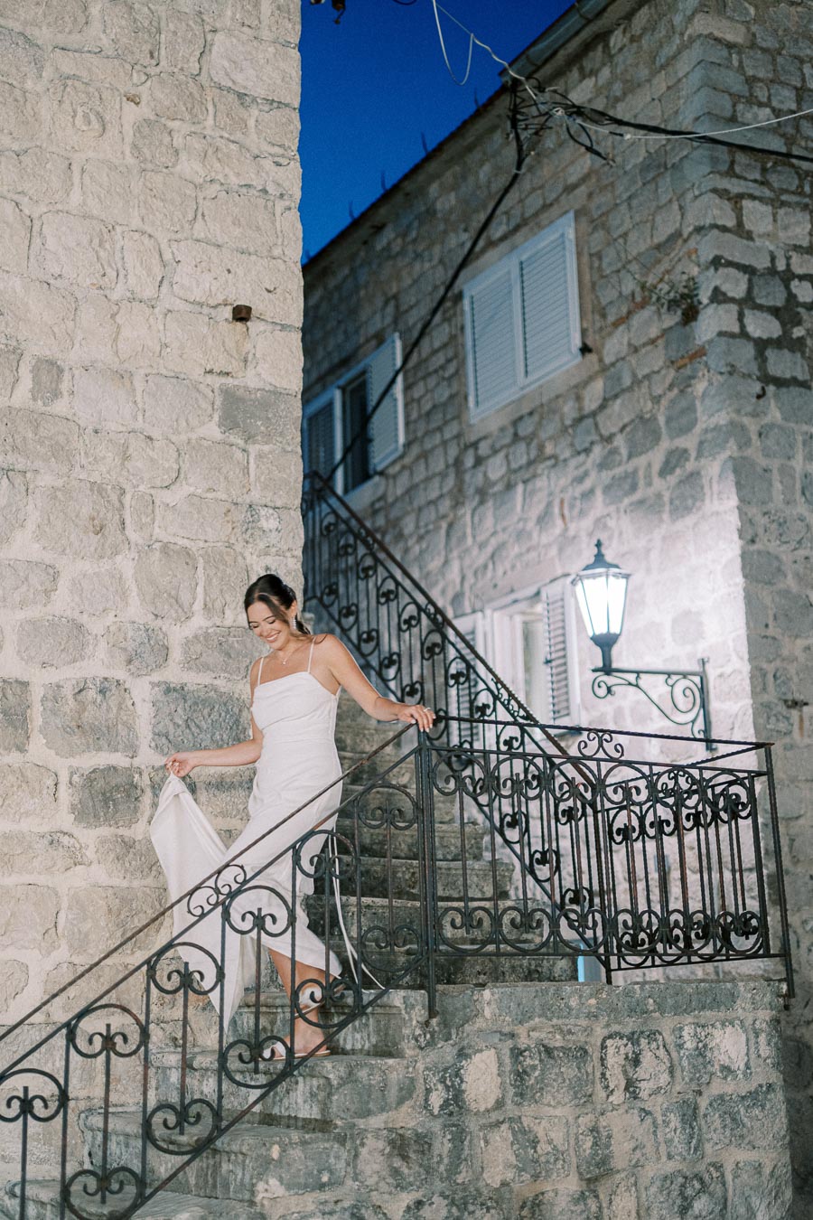 A bride in a white wedding dress descends an elegant wrought-iron staircase against the backdrop of an old stone building, illuminated by a soft streetlamp in a romantic evening setting.