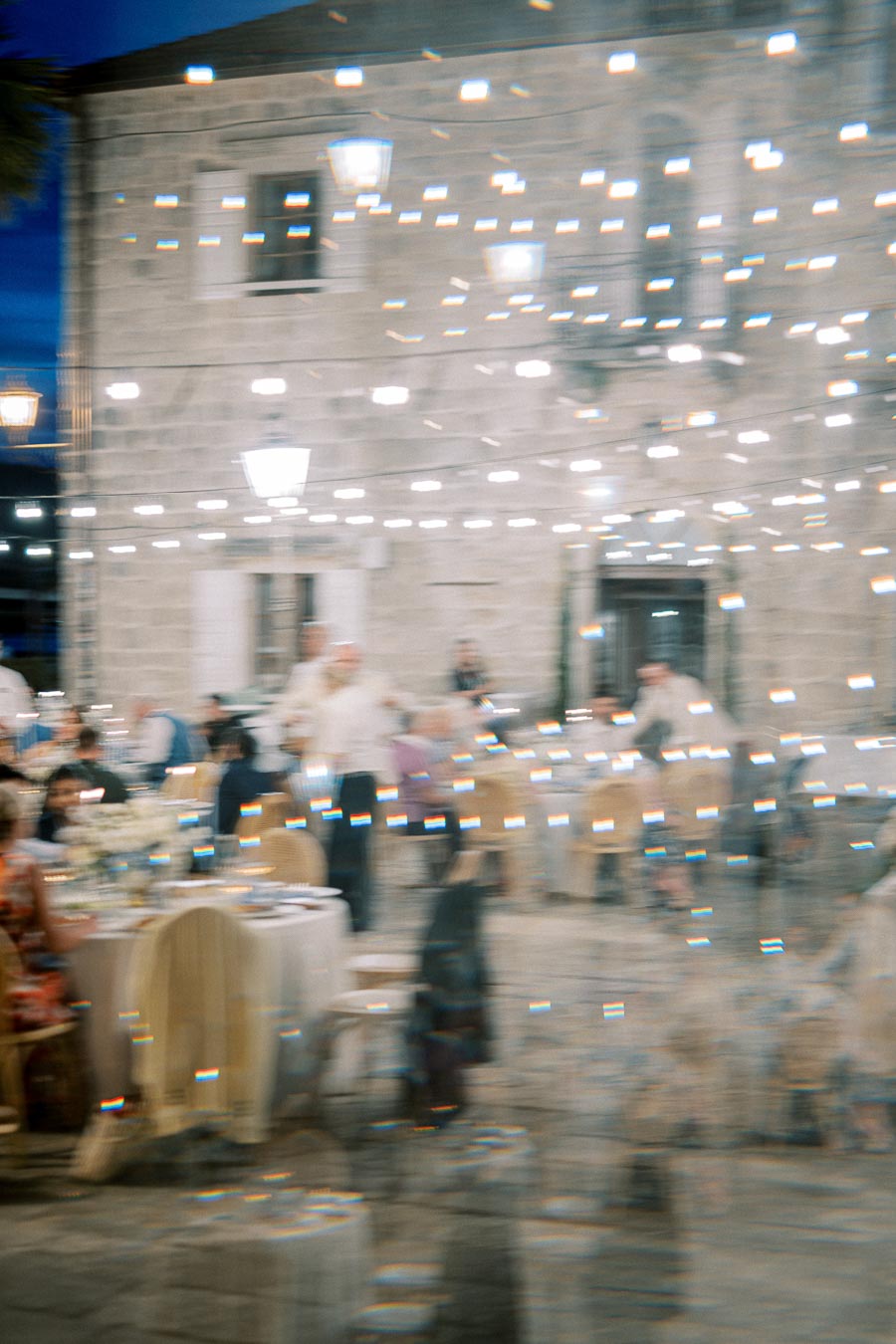 Blurred outdoor banquet scene with string lights illuminating guests seated at tables, set in front of an old stone building at night.