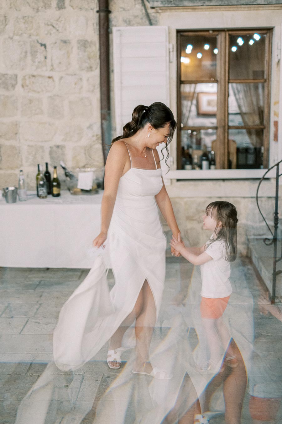 Smiling woman in a white dress joyfully dances with a young girl at an outdoor event, with a stone wall and window in the background.
