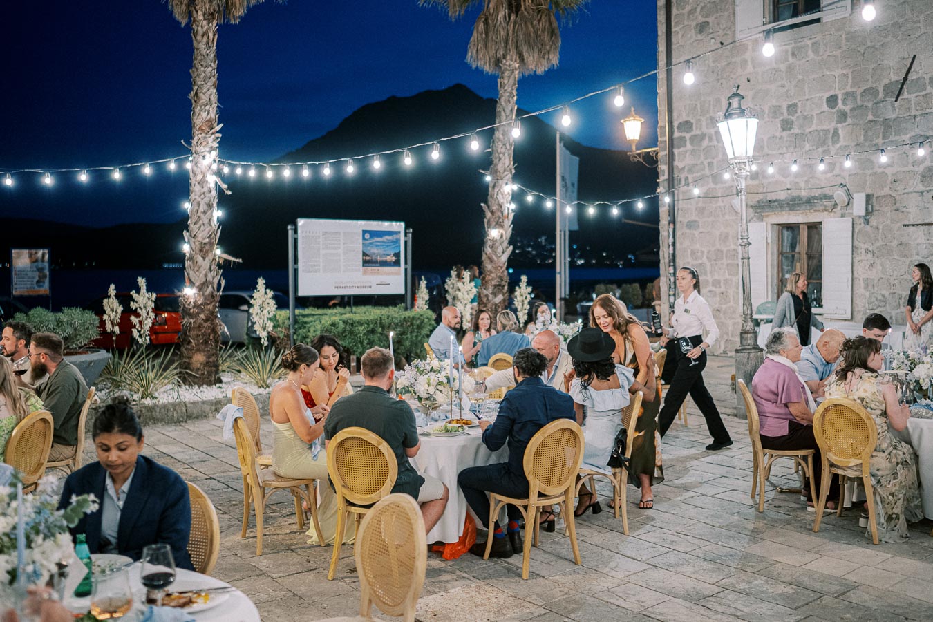 Outdoor evening event with people dining under string lights near the ocean, featuring elegantly set tables and stone architecture.