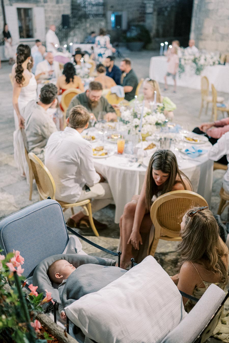 Outdoor wedding reception with elegantly dressed guests seated at tables adorned with white florals and candles, overlooking a cozy baby sleeping in a stroller surrounded by summer blooms.
