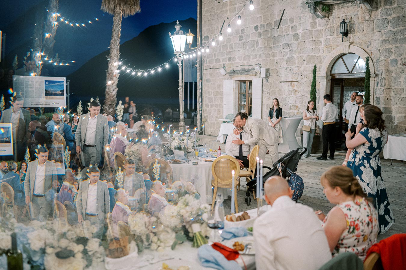Outdoor evening wedding reception with guests gathered around decorated tables under string lights, set against a rustic stone building.