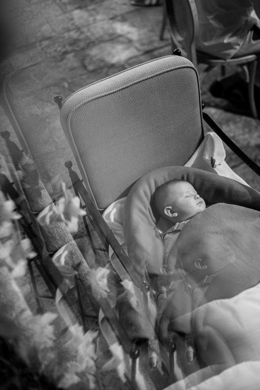 Black and white photograph of a sleeping baby in a stroller, with reflections creating a dreamy effect. The scene includes a patterned chair and blurred outdoor elements.