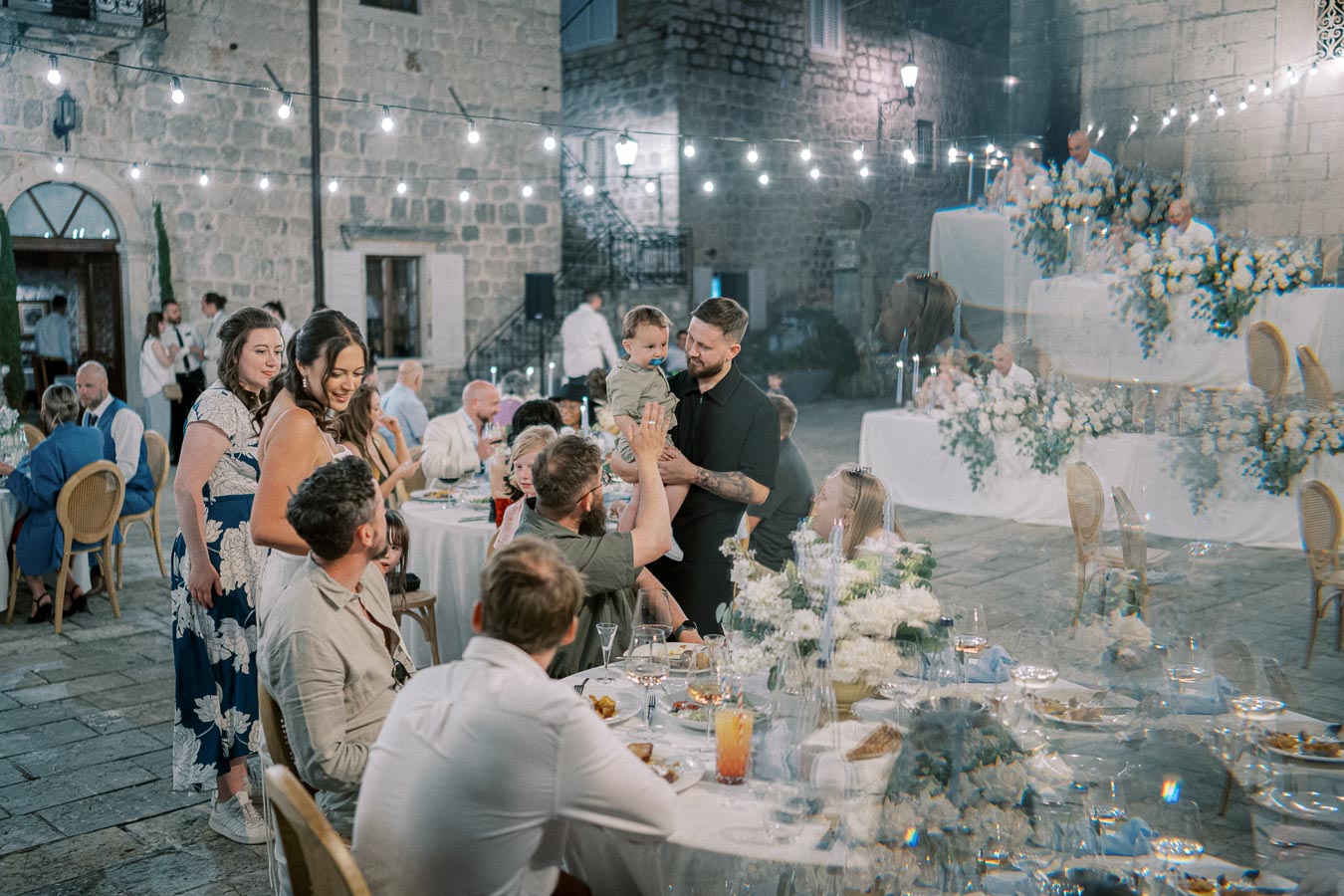 Outdoor wedding reception scene with guests gathered around tables adorned with white flowers and elegant place settings, under string lights. A man holds a toddler, surrounded by cheerful attendees. Romantic, rustic ambiance in a stone courtyard setting.