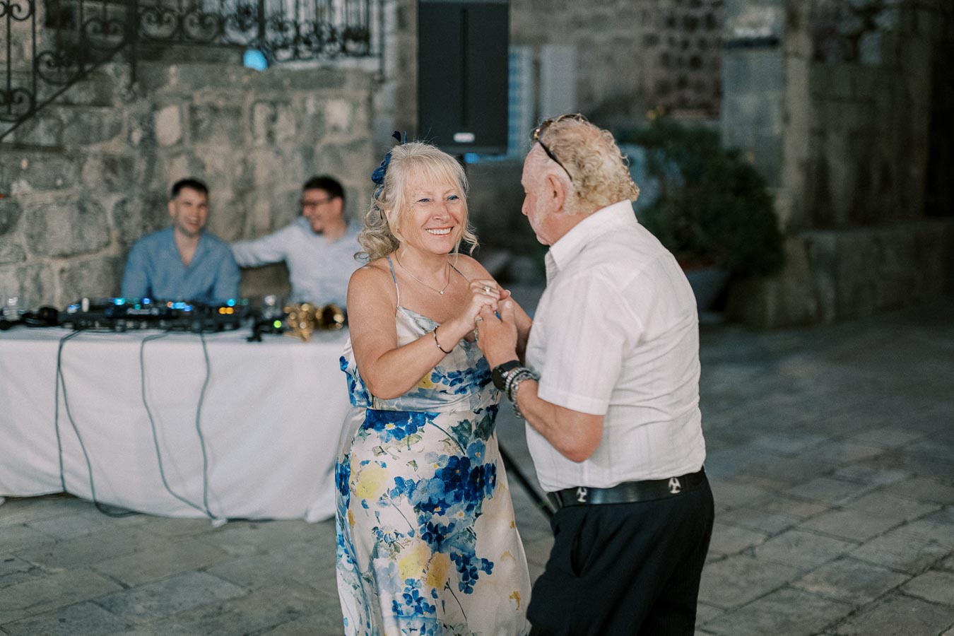 Elderly couple joyfully dancing at a wedding reception, with a DJ setup and stone wall in the background.