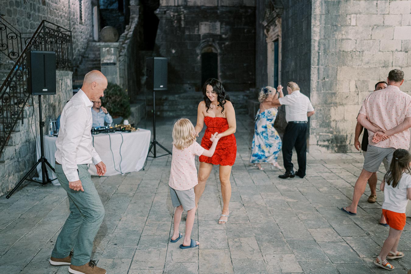Group of people dancing joyfully at an outdoor party in a historic stone courtyard, with a DJ setup in the background.