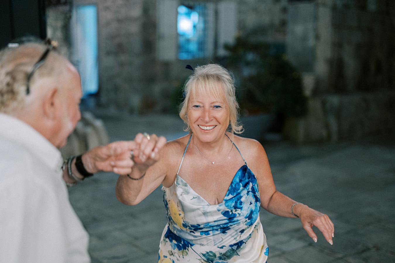 Elderly couple dancing joyfully in an outdoor setting, with the woman wearing a floral dress and smiling brightly, capturing a moment of happiness and companionship.