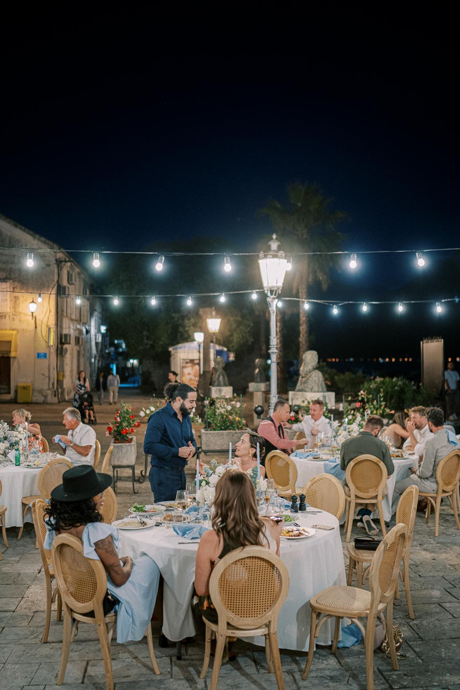 Outdoor evening dinner party with string lights and elegantly dressed guests seated at round tables.