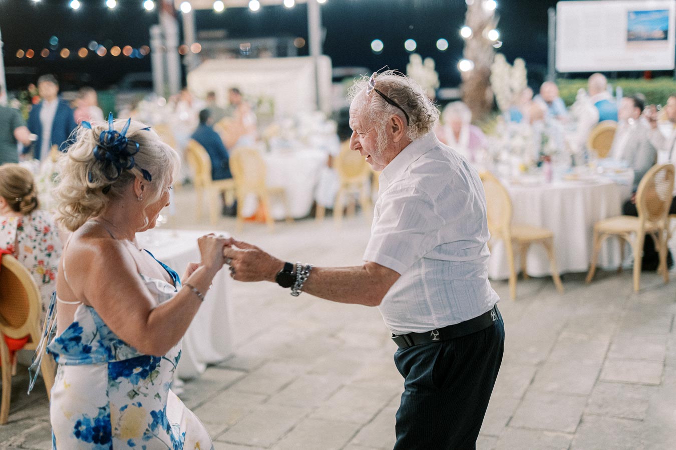 Elderly couple joyfully dancing at an outdoor evening celebration with guests seated at elegantly decorated tables in the background.