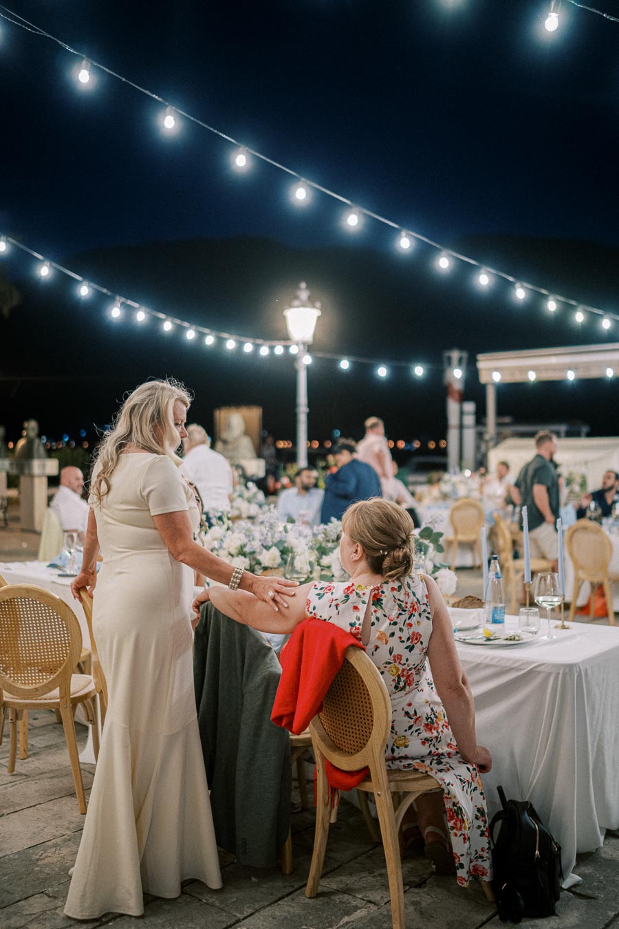 Elegant evening outdoor event with women in conversation under string lights, surrounded by decorated tables and guests.