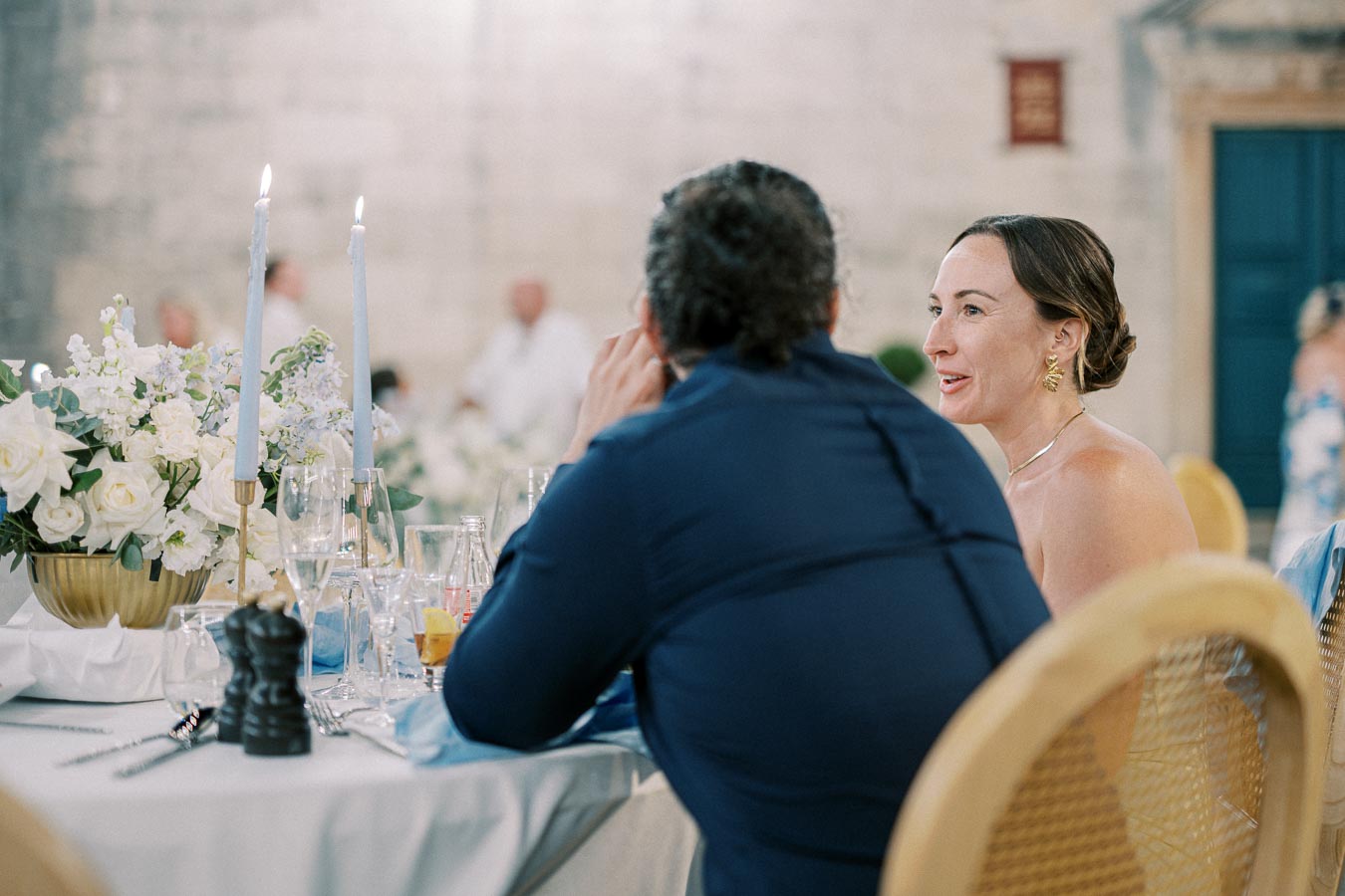 Elegant wedding reception with elegantly dressed guests seated at a beautifully decorated table adorned with white flowers and lit candles, creating a romantic atmosphere.