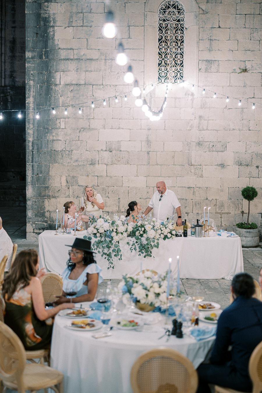 Elegant outdoor wedding reception scene with guests seated at round tables dressed in white tablecloths, adorned with floral centerpieces. The main table is decorated with lush greenery and white flowers, set against a stone wall with string lights hanging above, creating a romantic and festive atmosphere.