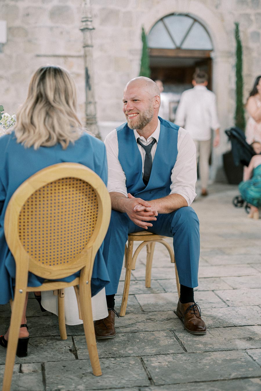 Smiling man in a blue vest and tie sits on a wooden chair, engaged in conversation at an outdoor event with stone walls in the background.
