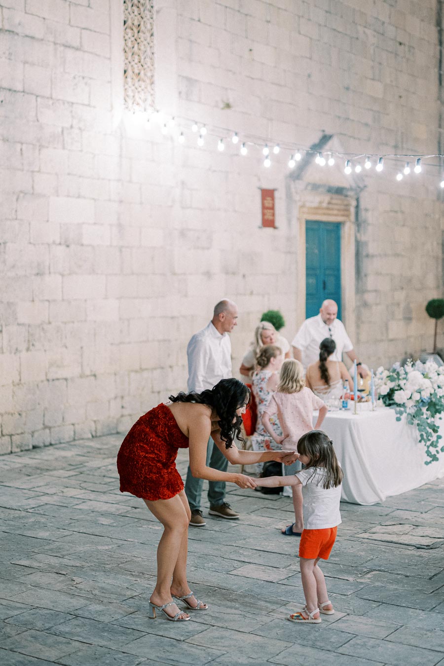 A woman in a red dress dances with a young girl at an outdoor event, surrounded by people and festive decorations. A stone wall and a table with a floral arrangement are visible in the background, creating a lively and joyful atmosphere.