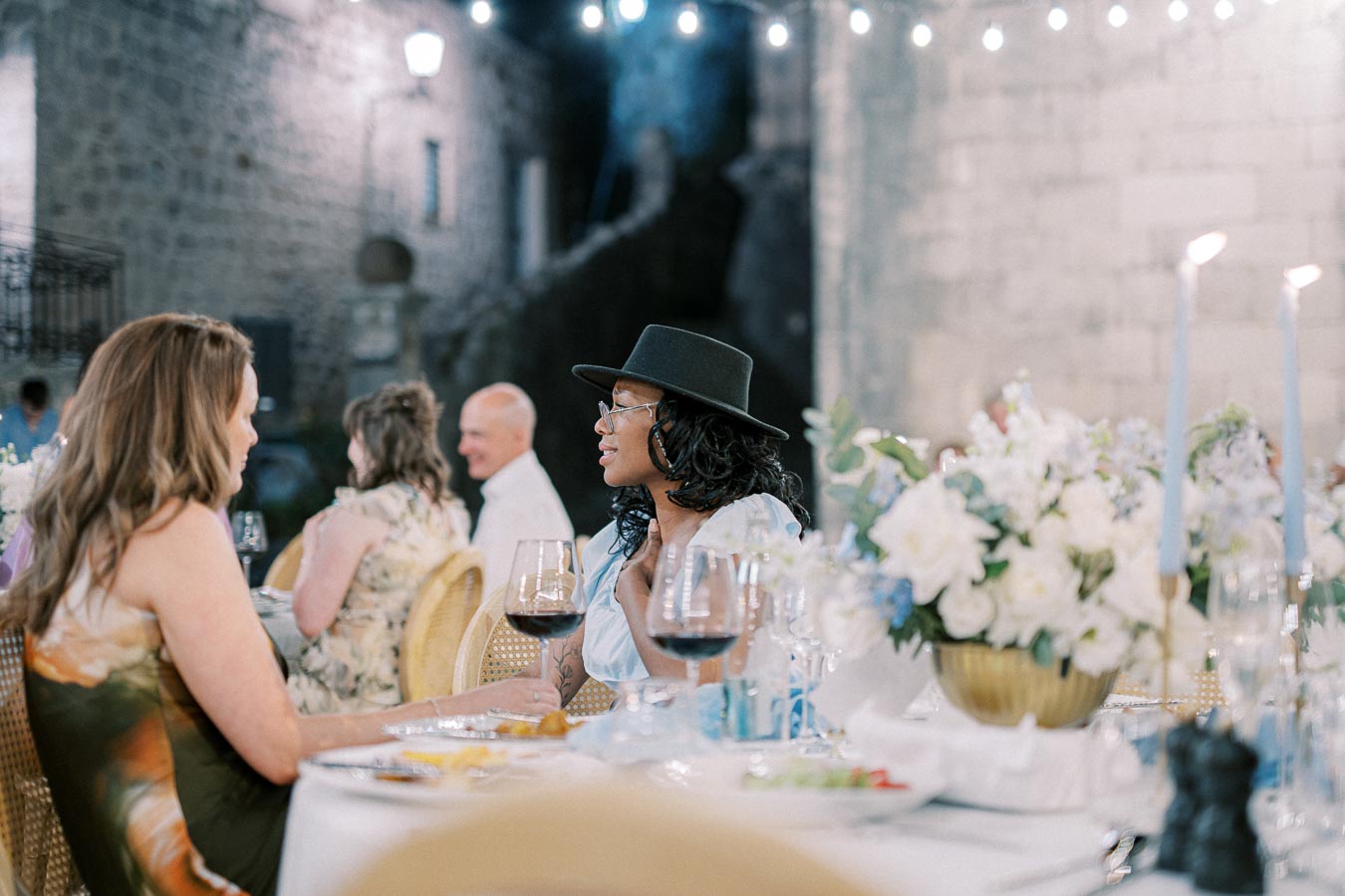 An elegant outdoor dinner event in a historic courtyard setting. Two women are engaged in conversation at a beautifully set table adorned with floral centerpieces and lit candles. Guests in the background enjoy the evening ambiance under string lights.
