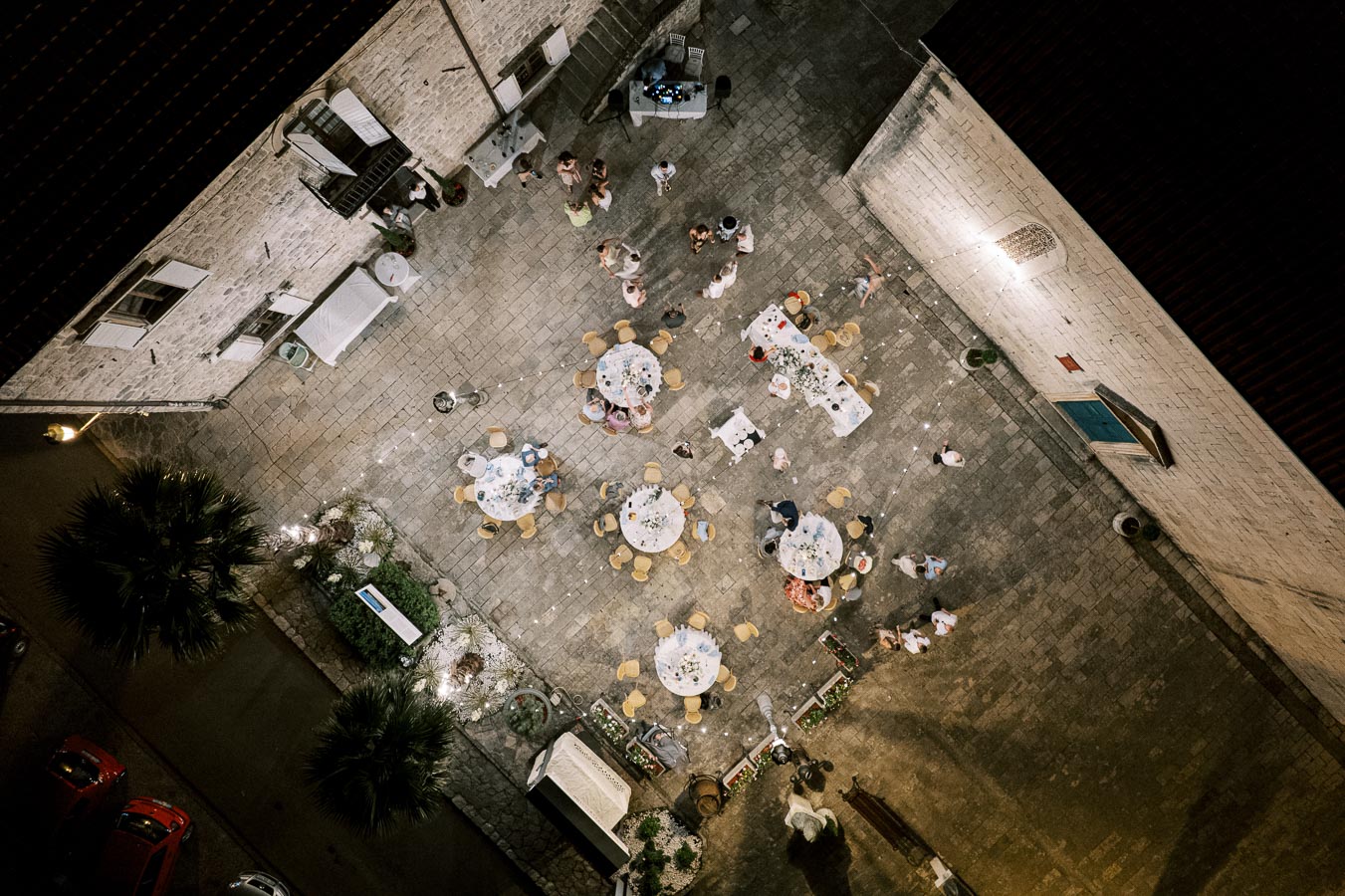 Aerial view of an outdoor evening event in a courtyard with round tables arranged for dining, surrounded by people socializing. An overhead string of lights illuminates the cobblestone area, creating a festive atmosphere.