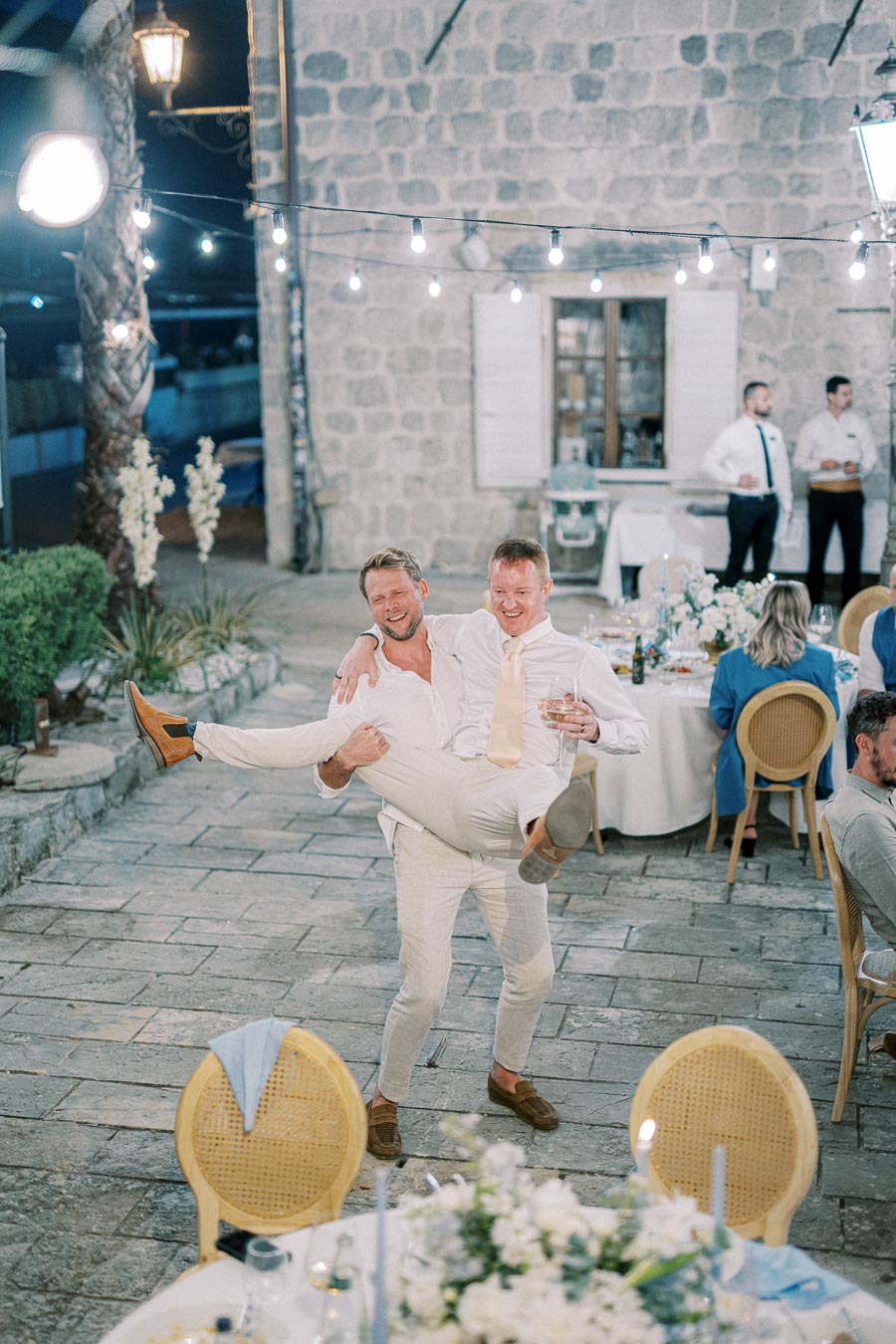 Two joyful men in white attire celebrating at an outdoor wedding reception, with one man playfully lifting the other. The scene features tables with floral decorations and guests enjoying the evening under string lights.
