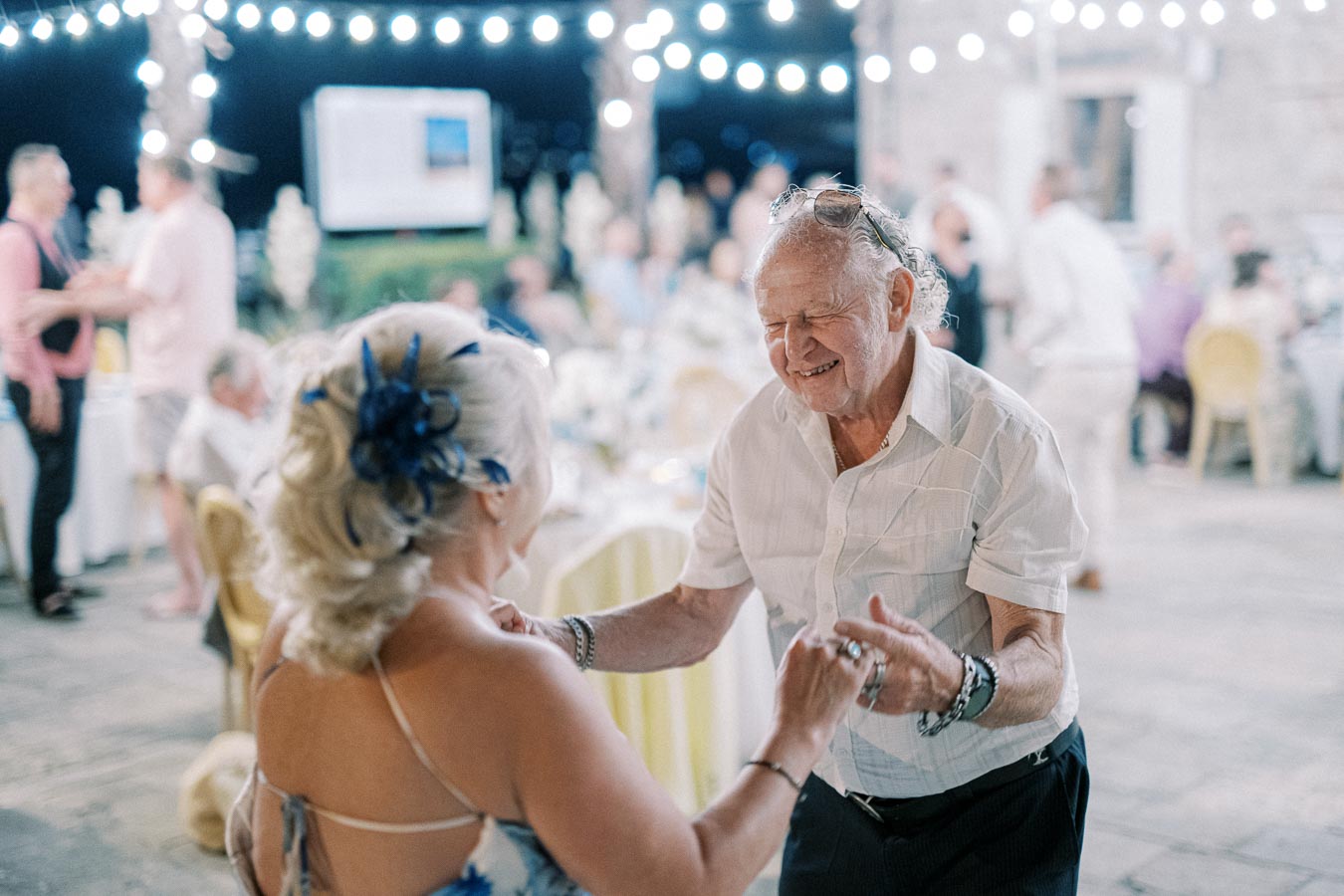 Elderly couple joyfully dancing at an outdoor evening event with decorative string lights, surrounded by guests and festive tables.